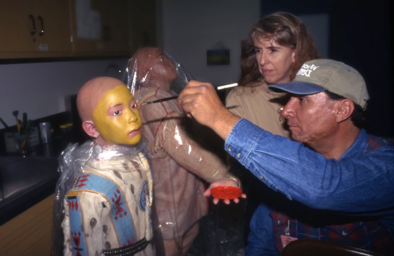 Allan painting a mannequin with the face paint for the weasel tail shirt for a Blackfoot display in the Royal Alberta Museum's former Gallery of Aboriginal Culture, 1997. Allan had the transferred rights to the face paint design as a weasel tail suit owner. (Photo Credit: Royal Alberta Museum)