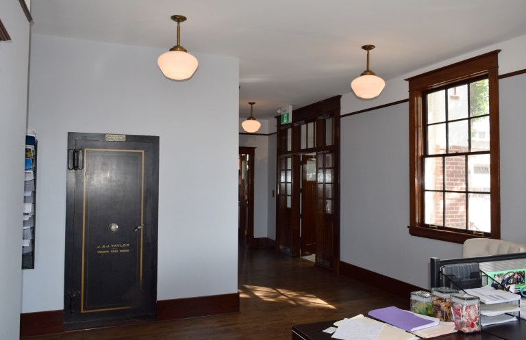 The main floor interior is currently an office after restoration of the damaged plaster walls and ceilings and conservation of the floors, windows and interior millwork. New pendant lights sourced by the owner feature “schoolhouse” globes that replicate the single historic fixture found in the original safe, at left. Source: Historic Resources Management Branch.